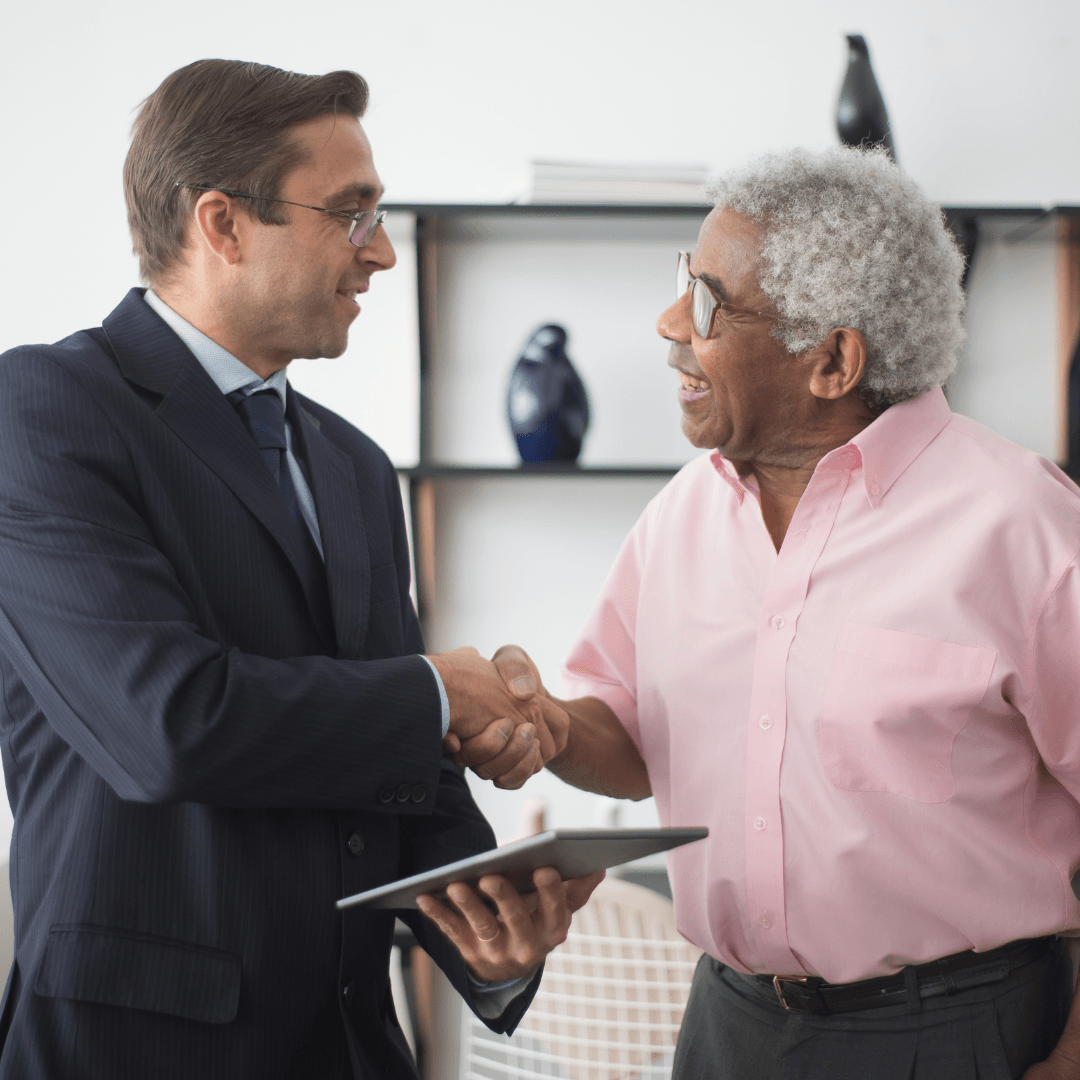 A suited financial professional wearing glasses shakes hands and smiles with an older man in a pink shirt during a meeting, with a tablet in hand.