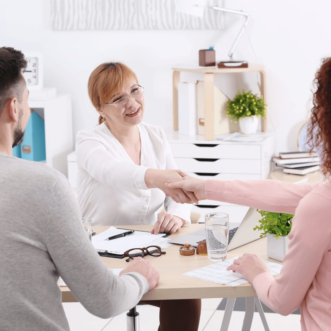 A smiling female advisor with blonde hair and glasses reaches across a desk to shake hands with a female client, while a male client sits beside her in a bright, modern office.