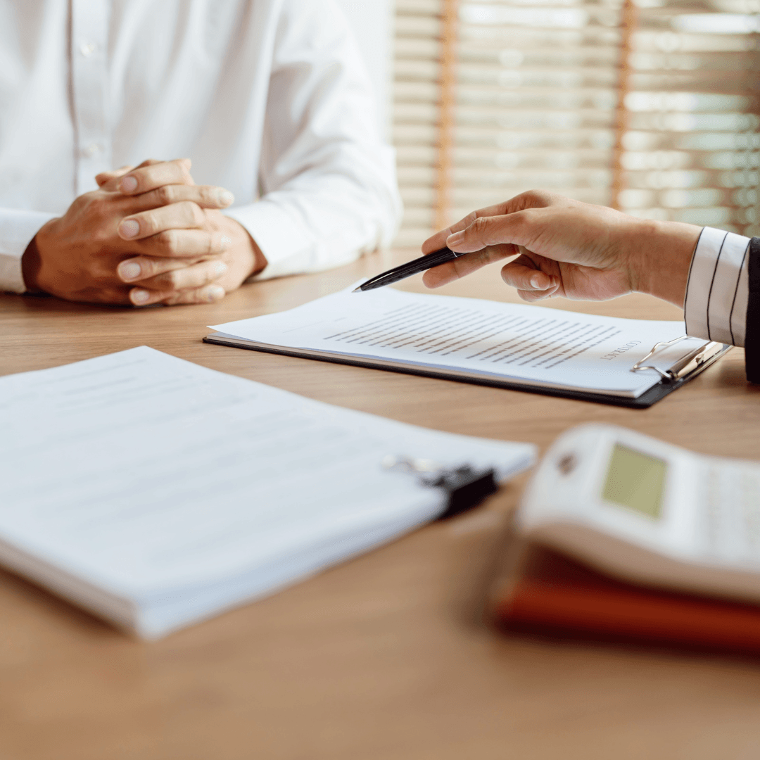 An advisor points a pen at a document on a clipboard across a wooden desk toward a client whose hands are clasped, with a second clipboard and calculator visible nearby.