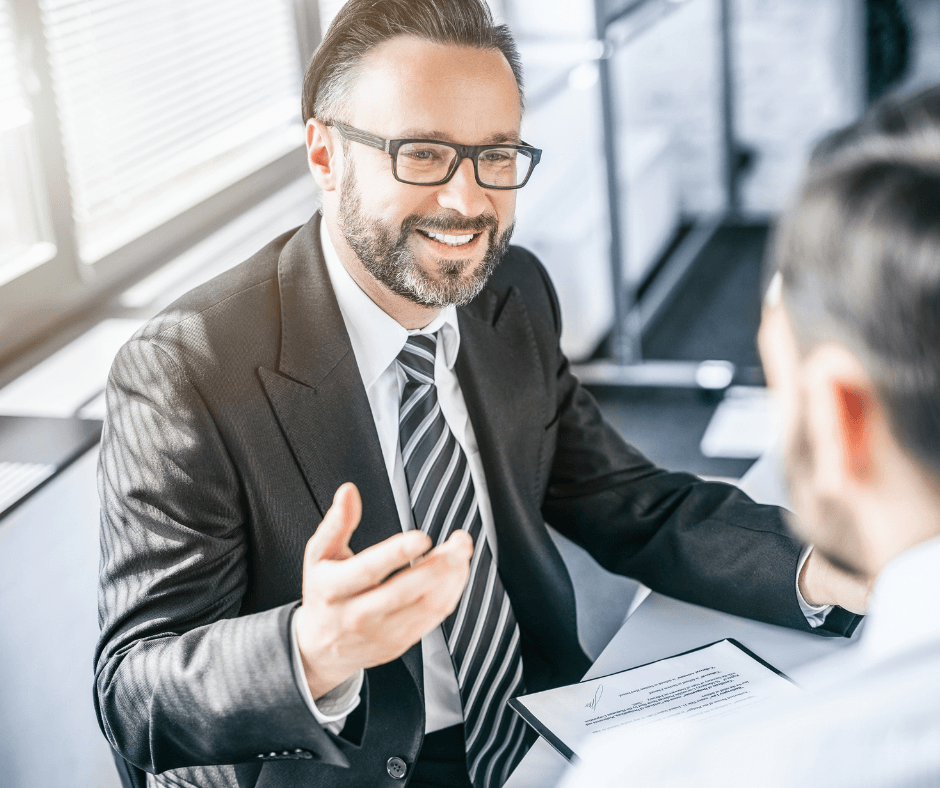 A smiling male insurance broker in a suit and glasses gestures while discussing a document with a client across a desk in a bright office setting.