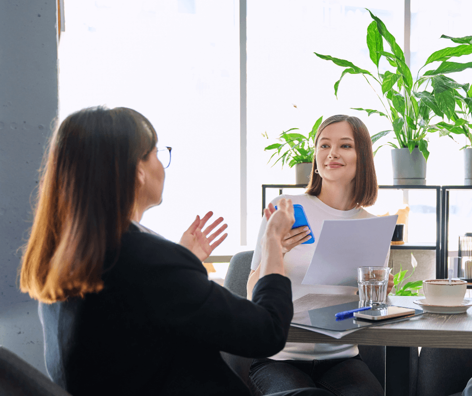 Two women sit across from each other at a table in a bright, plant-filled office space, reviewing documents during a consultation meeting.