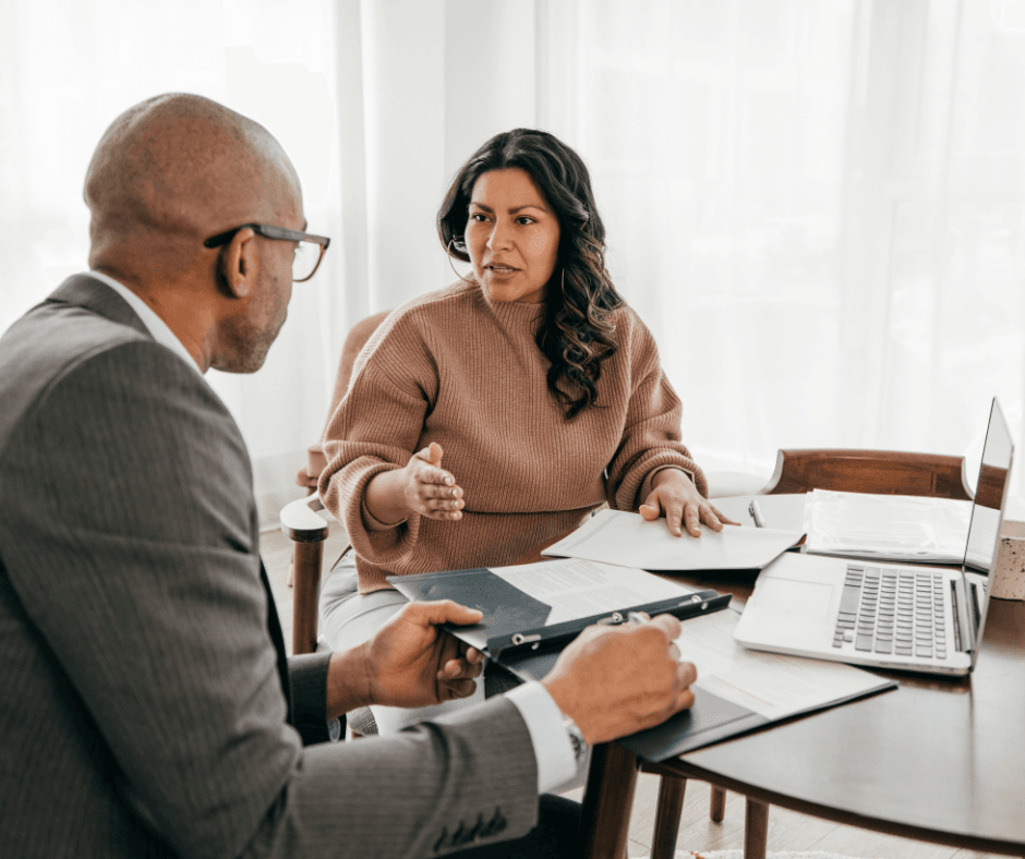 A woman in a rust-coloured sweater gestures while speaking with a male advisor holding a clipboard during an insurance consultation at a desk.