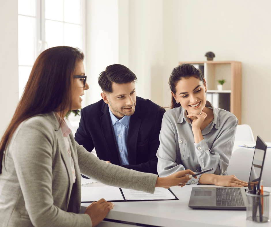 A young couple sits with a female advisor, all looking at a laptop screen together during a financial consultation meeting.