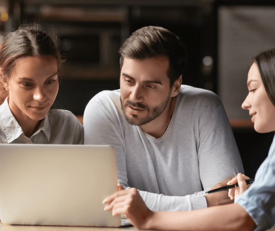 A female insurance advisor points to documents on a desk while a smiling couple reviews the information on a laptop during an office meeting.